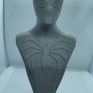 May include: A grey 3D printed bust of Spider-Man. The bust features a detailed sculpt of Spider-Man's head and torso, including the iconic spider symbol on his chest. The bust is mounted on a base with the words "SPIDER-MAN" printed on it.