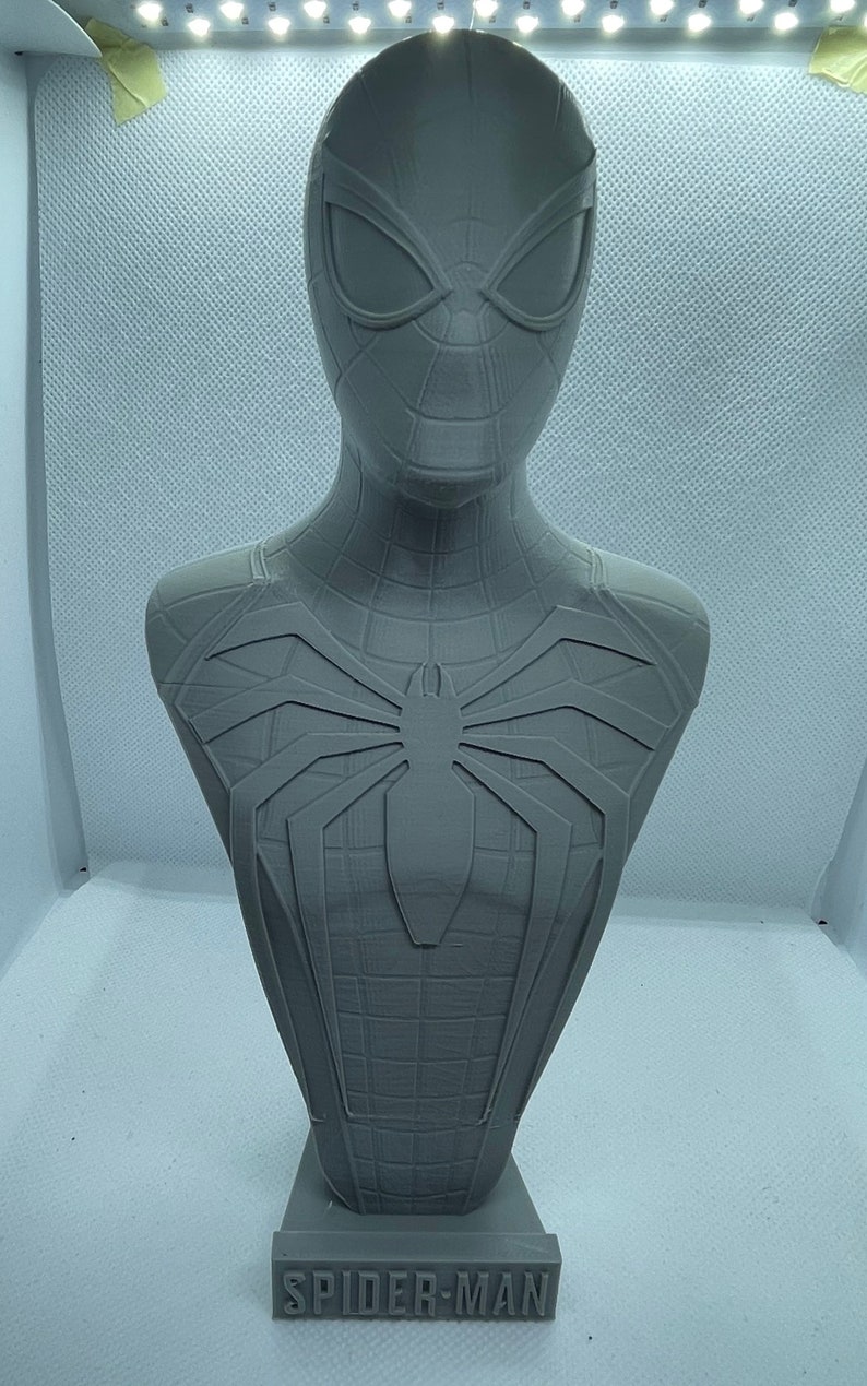 May include: A grey 3D printed bust of Spider-Man. The bust features a detailed sculpt of Spider-Man's head and torso, including the iconic spider symbol on his chest. The bust is mounted on a base with the words "SPIDER-MAN" printed on it.