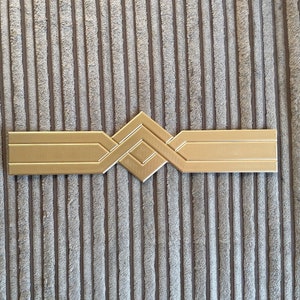 May include: A gold-coloured geometric design with a diamond shape in the centre. The design is made of three horizontal bands with angled edges.