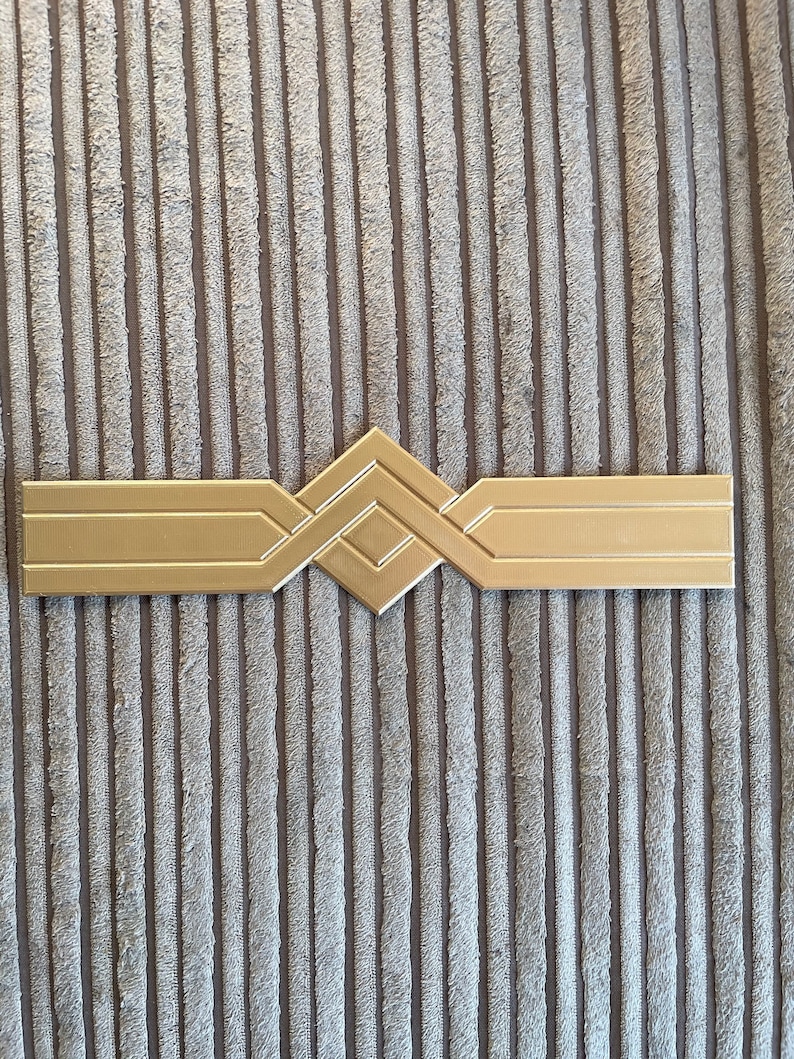 May include: A gold-coloured geometric design with a diamond shape in the centre. The design is made of three horizontal bands with angled edges.