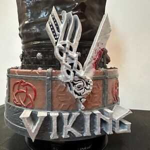 May include: A Viking-themed sculpture with the word "VIKING" in silver lettering. The sculpture includes a silver axe with red details, a shield, and a decorative knot design. The base is black.