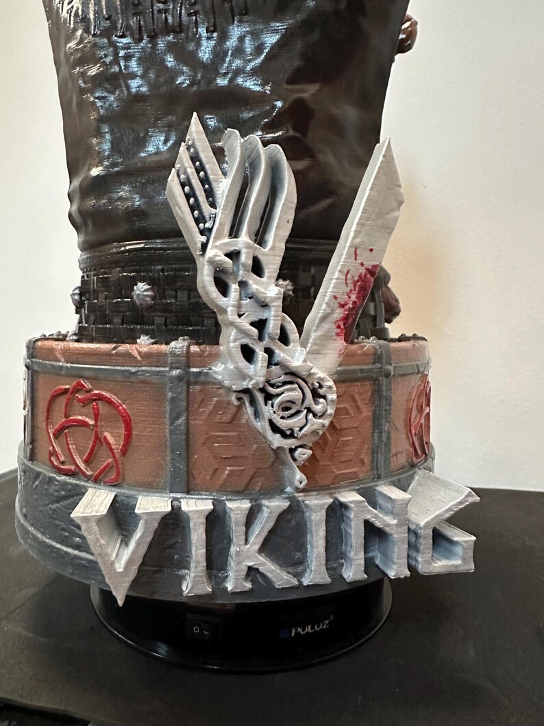 May include: A Viking-themed sculpture with the word "VIKING" in silver lettering. The sculpture includes a silver axe with red details, a shield, and a decorative knot design. The base is black.