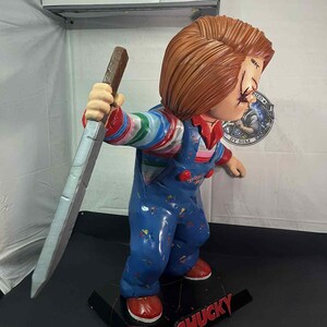May include: A Chucky figurine, featuring a red-haired doll in blue overalls, holding a large knife. The doll has stitched scars on its face and is standing on a black base with the name "CHUCKY" in red.