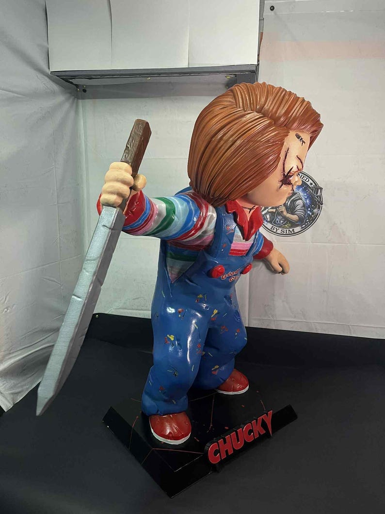 May include: A Chucky figurine, featuring a red-haired doll in blue overalls, holding a large knife. The doll has stitched scars on its face and is standing on a black base with the name "CHUCKY" in red.