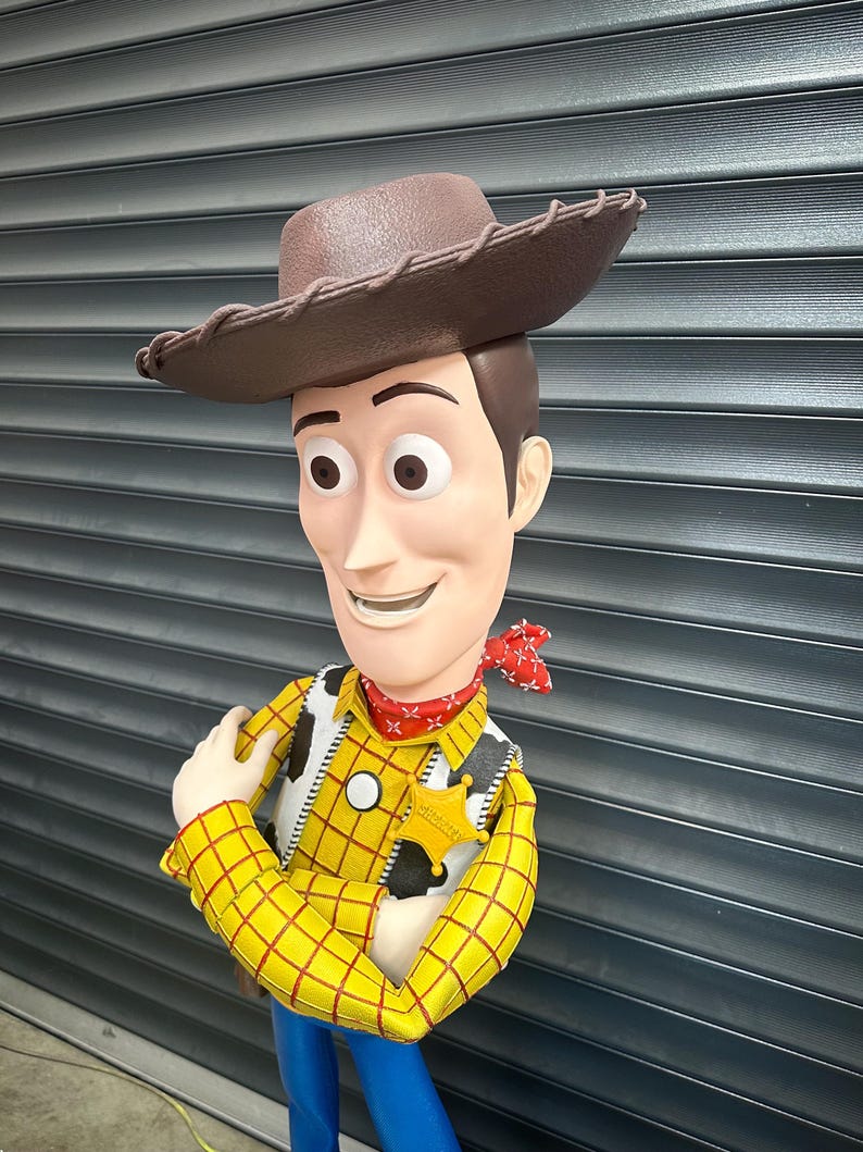 May include: A figurine of the character Woody from Toy Story. Woody is wearing a brown cowboy hat, a yellow plaid shirt, a vest, a red bandana, and blue trousers. He has a sheriff's badge and is smiling.