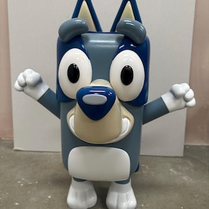 May include: A blue and white cartoon dog toy with a wide grin and large eyes. The toy is made of plastic and has a blocky shape.