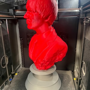 May include: A vibrant red bust of a man with a distinctive hairstyle and suit jacket. The sculpture is mounted on a gray, tiered base. The bust is likely a 3D-printed item, captured within a 3D printer.