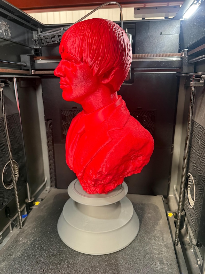 May include: A vibrant red bust of a man with a distinctive hairstyle and suit jacket. The sculpture is mounted on a gray, tiered base. The bust is likely a 3D-printed item, captured within a 3D printer.