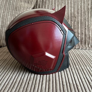 以下が含まれることがあります： テクスチャ加工された表面を持つ赤と黒のデアデビルヘルメット。ヘルメットは尖ったトップと黒いマスクを持っています。
