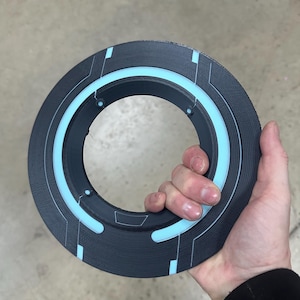 May include: A circular, black object with a light blue inner ring and geometric accents. The object is held in a hand, showcasing its design. The design features a series of straight lines and rectangular shapes, creating a futuristic aesthetic.