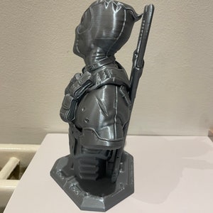 May include: A gray 3D printed bust of Deadpool, a superhero character, with a detailed design and a textured finish. The bust is posed with one arm raised and a weapon on his back.