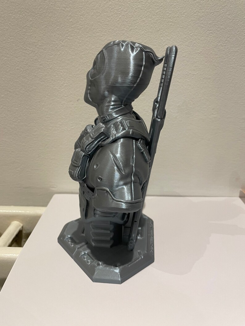 May include: A gray 3D printed bust of Deadpool, a superhero character, with a detailed design and a textured finish. The bust is posed with one arm raised and a weapon on his back.
