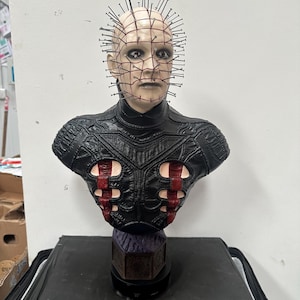 May include: A detailed bust of a character with a pale complexion, adorned with numerous metal pins protruding from the head. The figure wears a black leather-like garment with red accents. The bust is mounted on a dark base.