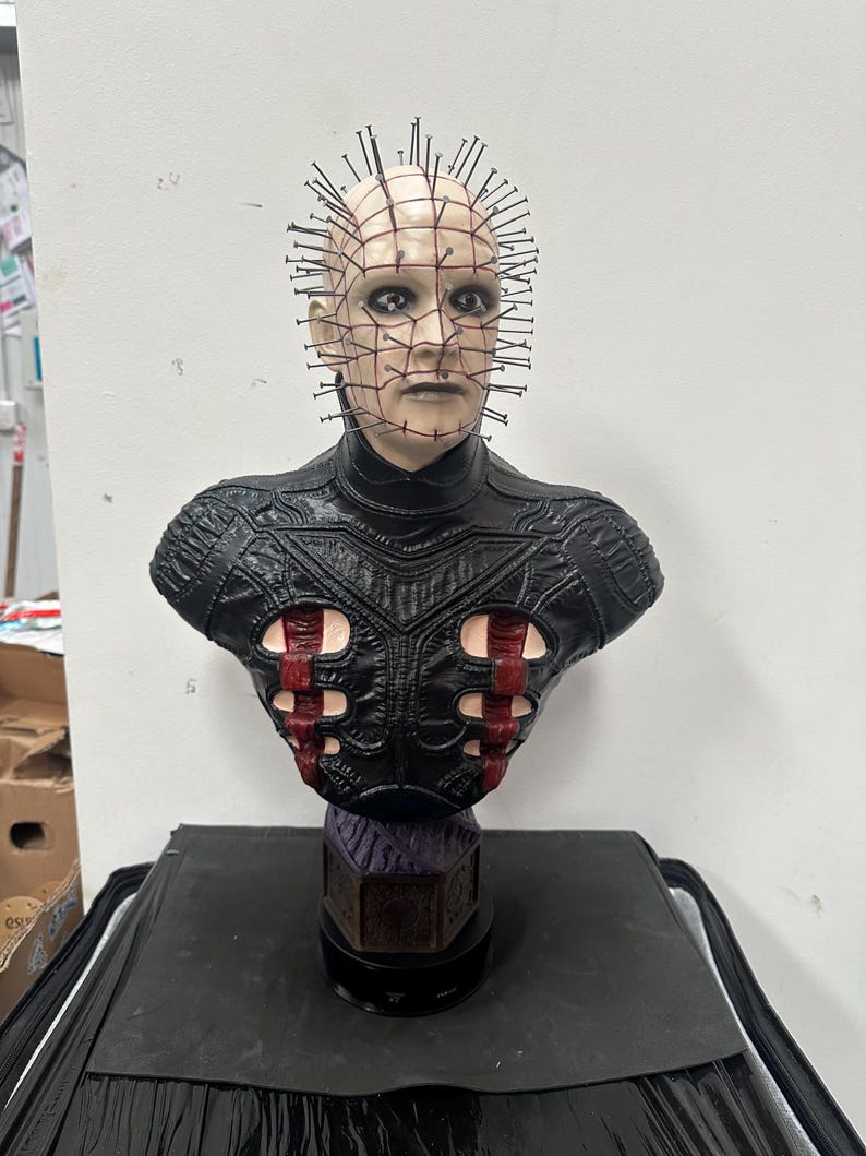 May include: A detailed bust of a character with a pale complexion, adorned with numerous metal pins protruding from the head. The figure wears a black leather-like garment with red accents. The bust is mounted on a dark base.