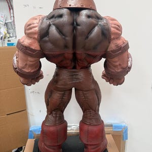May include: A detailed, rear view of a large, muscular action figure. The figure is predominantly brown and red, with a textured appearance. It features a helmet, large shoulders, and boots. The figure is posed on a black surface.