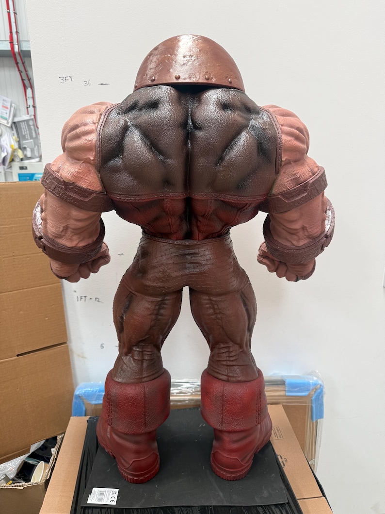 May include: A detailed, rear view of a large, muscular action figure. The figure is predominantly brown and red, with a textured appearance. It features a helmet, large shoulders, and boots. The figure is posed on a black surface.