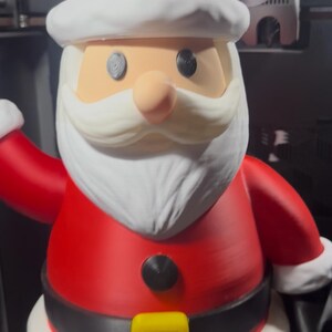 May include: A 3D-printed Santa Claus figurine. The figure is predominantly red with white trim on the hat, sleeves, and base. It has a white beard, a peach-colored nose, and black eyes. A black belt with a yellow buckle is visible.