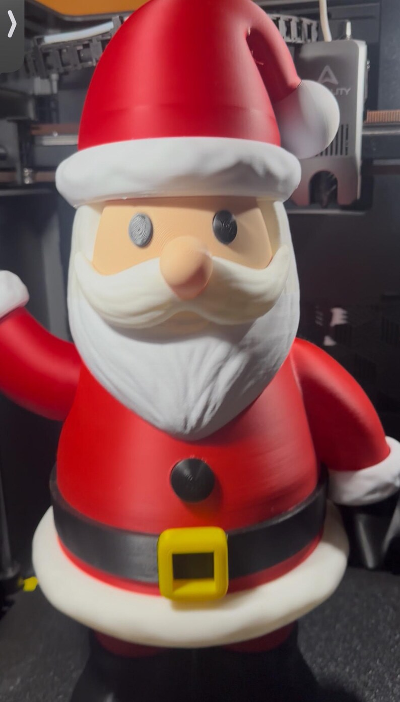May include: A 3D-printed Santa Claus figurine. The figure is predominantly red with white trim on the hat, sleeves, and base. It has a white beard, a peach-colored nose, and black eyes. A black belt with a yellow buckle is visible.