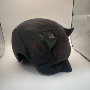 May include: A black helmet with a textured surface and two small horns. The helmet has red accents and a curved visor. The design suggests a superhero or cosplay theme.