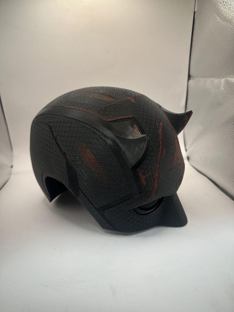 May include: A black helmet with a textured surface and two small horns. The helmet has red accents and a curved visor. The design suggests a superhero or cosplay theme.