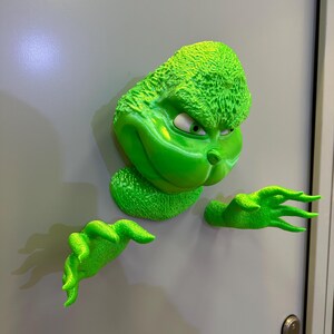 May include: A green Grinch character figurine with a mischievous expression, peeking around a door. The figurine is 3D printed and has a textured surface.