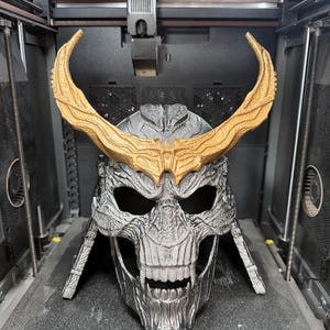 May include: A 3D printed skull-shaped helmet featuring a metallic silver finish and large, textured gold-colored horns extending from the forehead. The helmet displays intricate bone-like detailing, deep eye sockets, and a bared-teeth jaw structure.