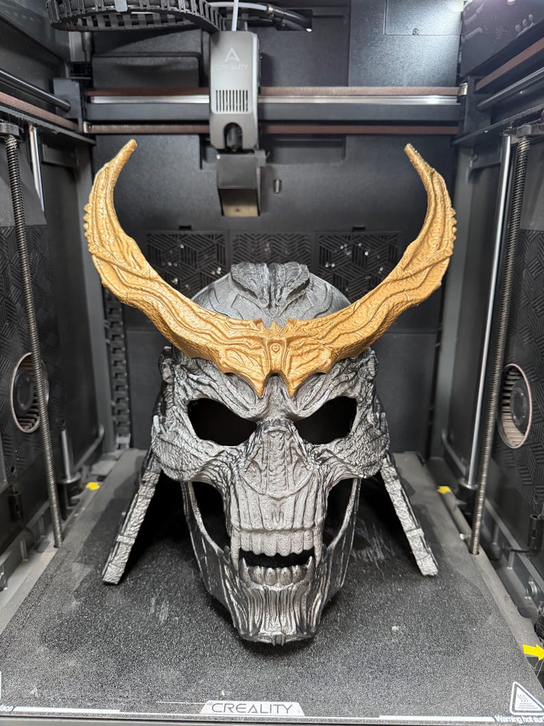 May include: A 3D printed skull-shaped helmet featuring a metallic silver finish and large, textured gold-colored horns extending from the forehead. The helmet displays intricate bone-like detailing, deep eye sockets, and a bared-teeth jaw structure.