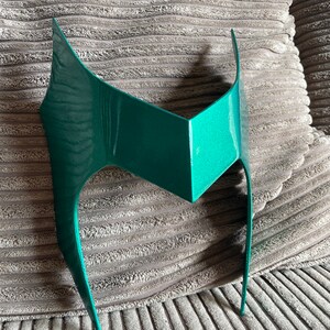 May include: A teal-colored, angular mask with a pointed top and curved sides. The mask has a metallic sheen and is designed to be worn on the forehead. The mask is likely for costume or cosplay purposes.