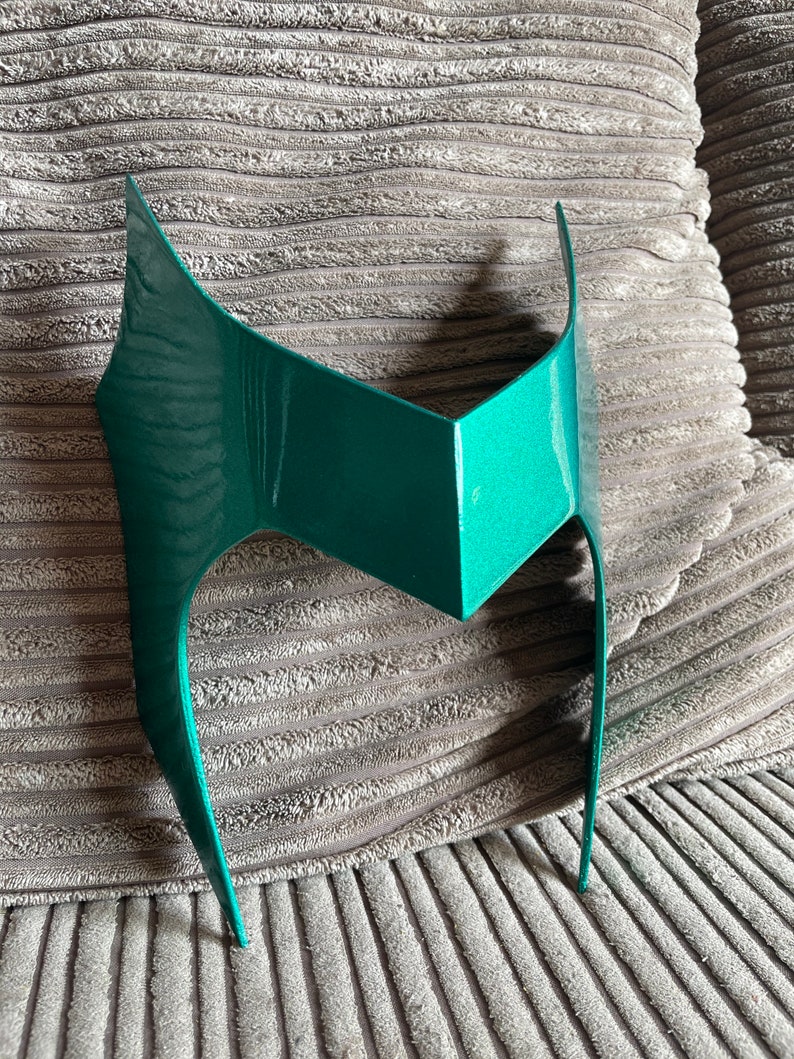 May include: A teal-colored, angular mask with a pointed top and curved sides. The mask has a metallic sheen and is designed to be worn on the forehead. The mask is likely for costume or cosplay purposes.