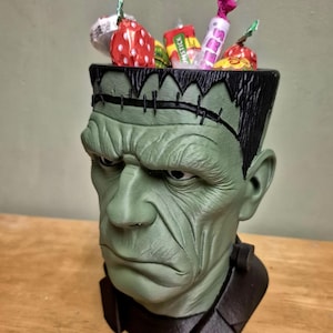 Puede incluir: Un recipiente verde con forma de cabeza de Frankenstein lleno de caramelos envueltos. El recipiente tiene cabello negro y detalles de costura. Los caramelos son de varios colores y formas, incluyendo rojo, blanco y amarillo.