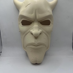 May include: A cream-colored mask with a stern expression and prominent features. The mask has large, curved horns, deep eye sockets, and a defined nose and mouth. The surface has a textured appearance, with a crack on the forehead. A unique decorative piece.