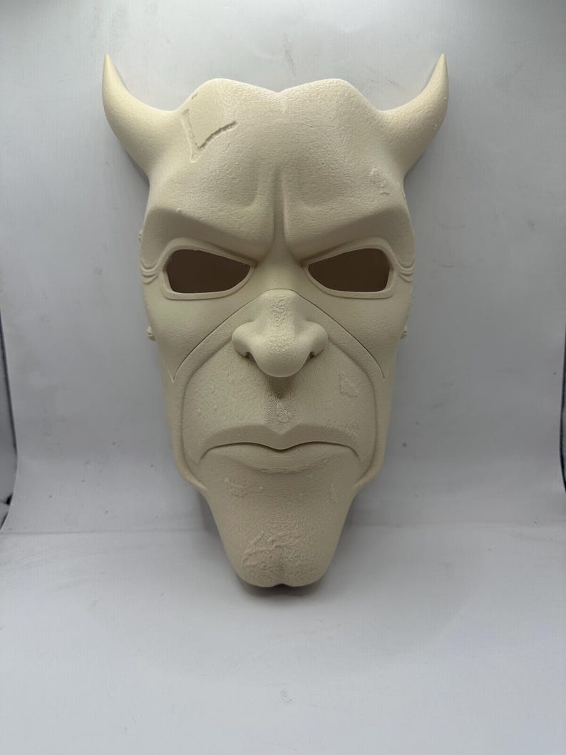 May include: A cream-colored mask with a stern expression and prominent features. The mask has large, curved horns, deep eye sockets, and a defined nose and mouth. The surface has a textured appearance, with a crack on the forehead. A unique decorative piece.
