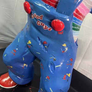 May include: A blue figurine of a character wearing overalls with the text "Good Guys" in red. The overalls are decorated with small tool graphics. The figurine has red shoes and red buttons on the overalls.