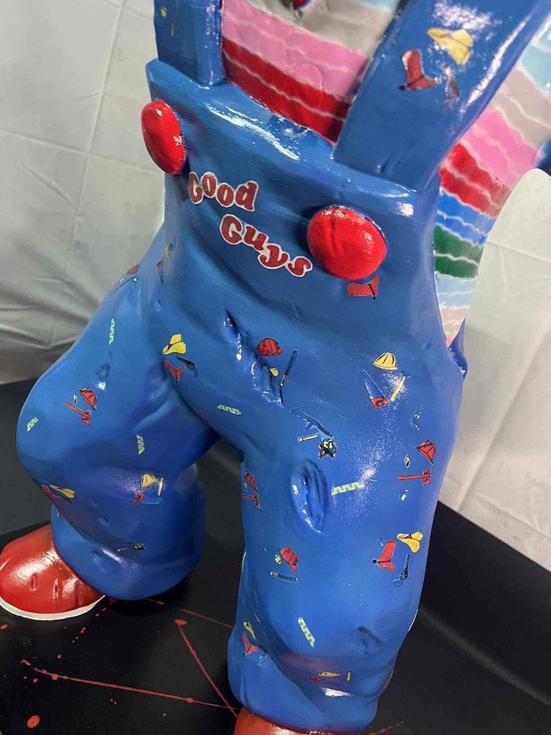 May include: A blue figurine of a character wearing overalls with the text "Good Guys" in red. The overalls are decorated with small tool graphics. The figurine has red shoes and red buttons on the overalls.