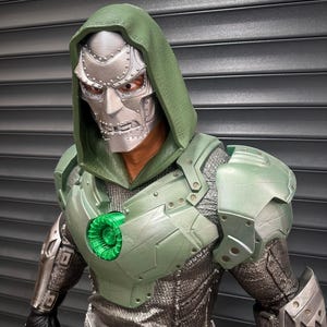 May include: A detailed action figure of a character with a silver mask, green hood, and armor. The figure has a metallic appearance with intricate details on the mask and armor. The chest piece features a glowing green circular element. The figure is posed against a gray background.