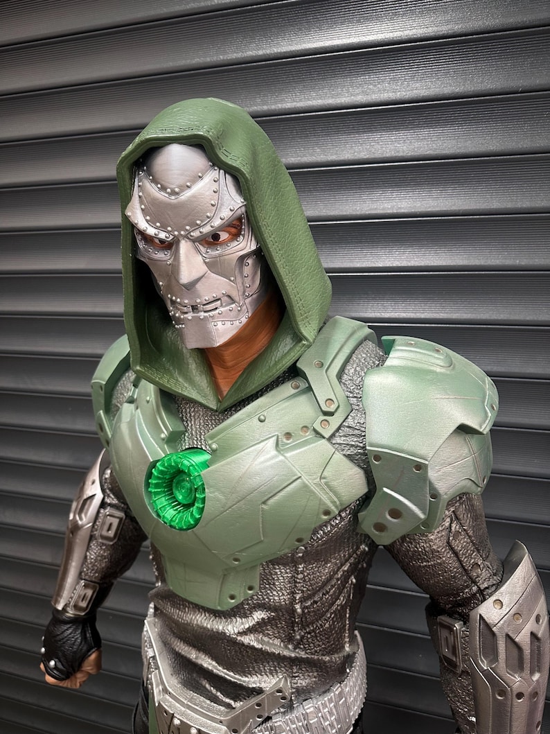 May include: A detailed action figure of a character with a silver mask, green hood, and armor. The figure has a metallic appearance with intricate details on the mask and armor. The chest piece features a glowing green circular element. The figure is posed against a gray background.