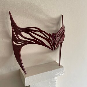 May include: A burgundy-colored, intricate, decorative mask made of plastic. The mask is shaped like a crown and has a delicate, lacy design.