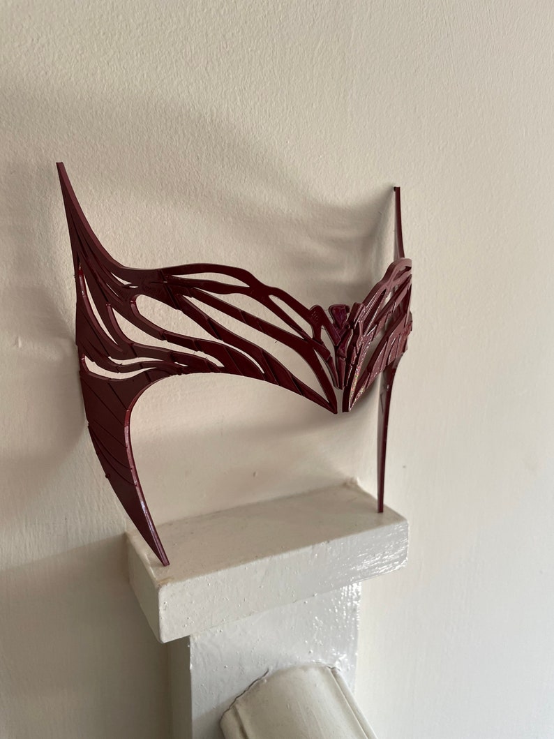 May include: A burgundy-colored, intricate, decorative mask made of plastic. The mask is shaped like a crown and has a delicate, lacy design.