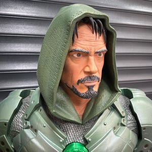 May include: A detailed action figure of a man in a green hooded costume with armor. The figure has a stern expression, a goatee, and a green chest piece with a glowing green center. The armor has a metallic finish.