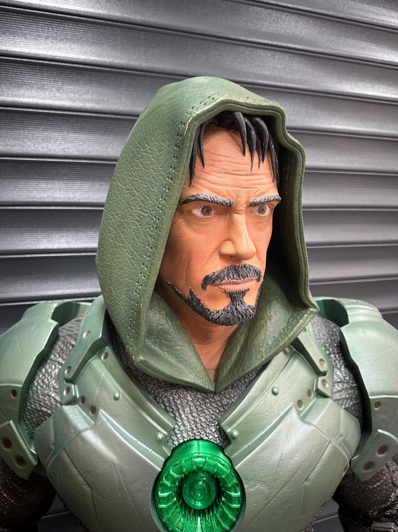 May include: A detailed action figure of a man in a green hooded costume with armor. The figure has a stern expression, a goatee, and a green chest piece with a glowing green center. The armor has a metallic finish.
