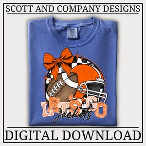 May include: A periwinkle blue t-shirt featuring a graphic of a brown football, an orange football helmet, and a large orange bow. The design includes a black and white checkered circle and the orange text "LET'S GO" with "jackets" in black script.