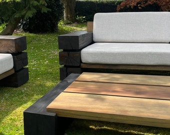 Puede incluir: Juego de muebles de jardín de madera con un sofá, dos sillones y una mesa de centro. El sofá y los sillones tienen cojines de color gris claro. Los marcos de madera están teñidos de negro y madera natural. El conjunto está sobre césped.