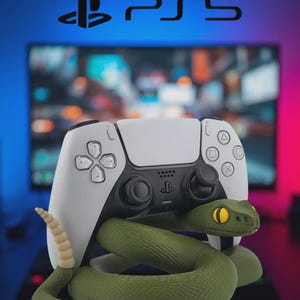 May include: A white and black wireless game controller resting on a green rattlesnake-shaped stand. The snake has a textured body, yellow eyes, and a tan rattle at the end of its tail. The PS5 logo is visible in the background above the controller.