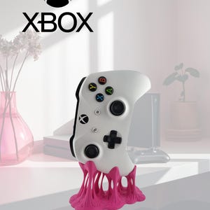 May include: A white Xbox wireless controller with black joysticks and colorful action buttons, resting on a bright pink, organic-shaped, textured stand. The Xbox logo and text are visible at the top of the image in black.