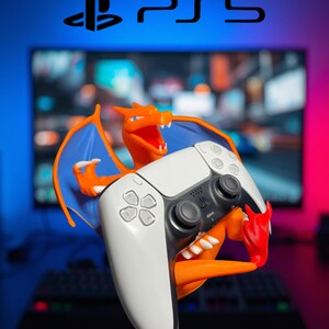 May include: An orange Charizard figure holding a white and black PS5 wireless controller. The figure features blue wings, sharp claws, and an open mouth. The PS5 logo is visible at the top. The controller has standard buttons, joysticks, and a touchpad.