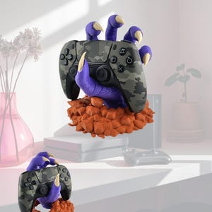 May include: A purple monster hand with sharp cream-colored claws emerging from a pile of orange rocks, holding a camouflage-patterned video game controller. The controller features black buttons, joysticks, and standard gaming symbols on the face.