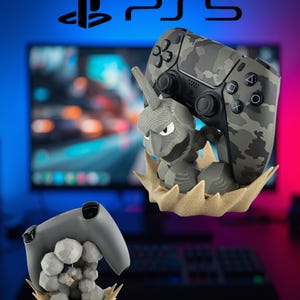May include: A gray and black camouflage PS5 controller resting on a textured gray rock-like figure stand with a tan base. A second view shows the back of a solid gray controller supported by a cluster of gray geometric rocks on a tan base.
