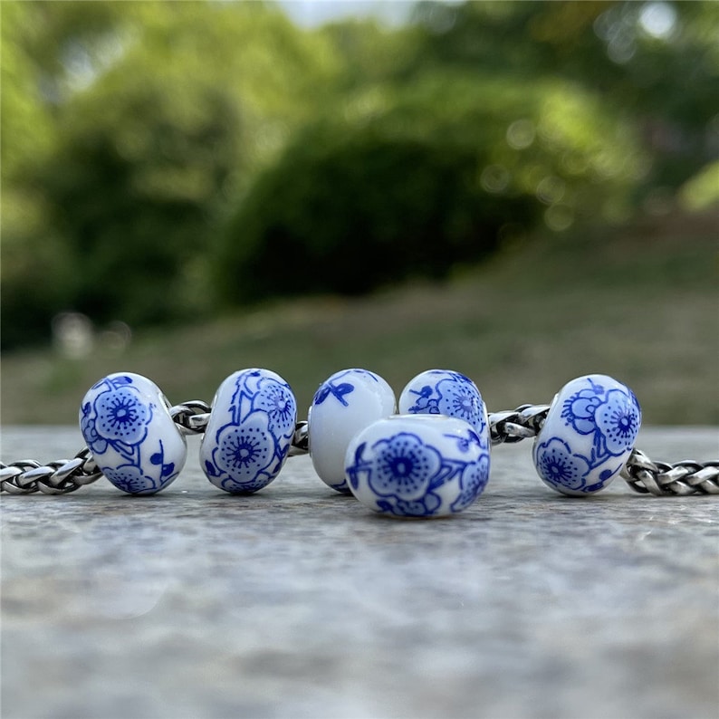 May include: Five white ceramic beads with blue floral patterns. The beads are arranged in a row on a wooden surface.
