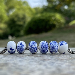 May include: Six white ceramic beads with blue floral patterns. The beads are strung on a silver chain.