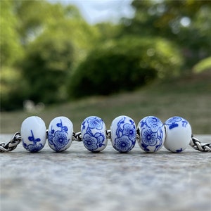 May include: Six white ceramic beads with blue floral patterns. The beads are arranged in a row on a brown surface.
